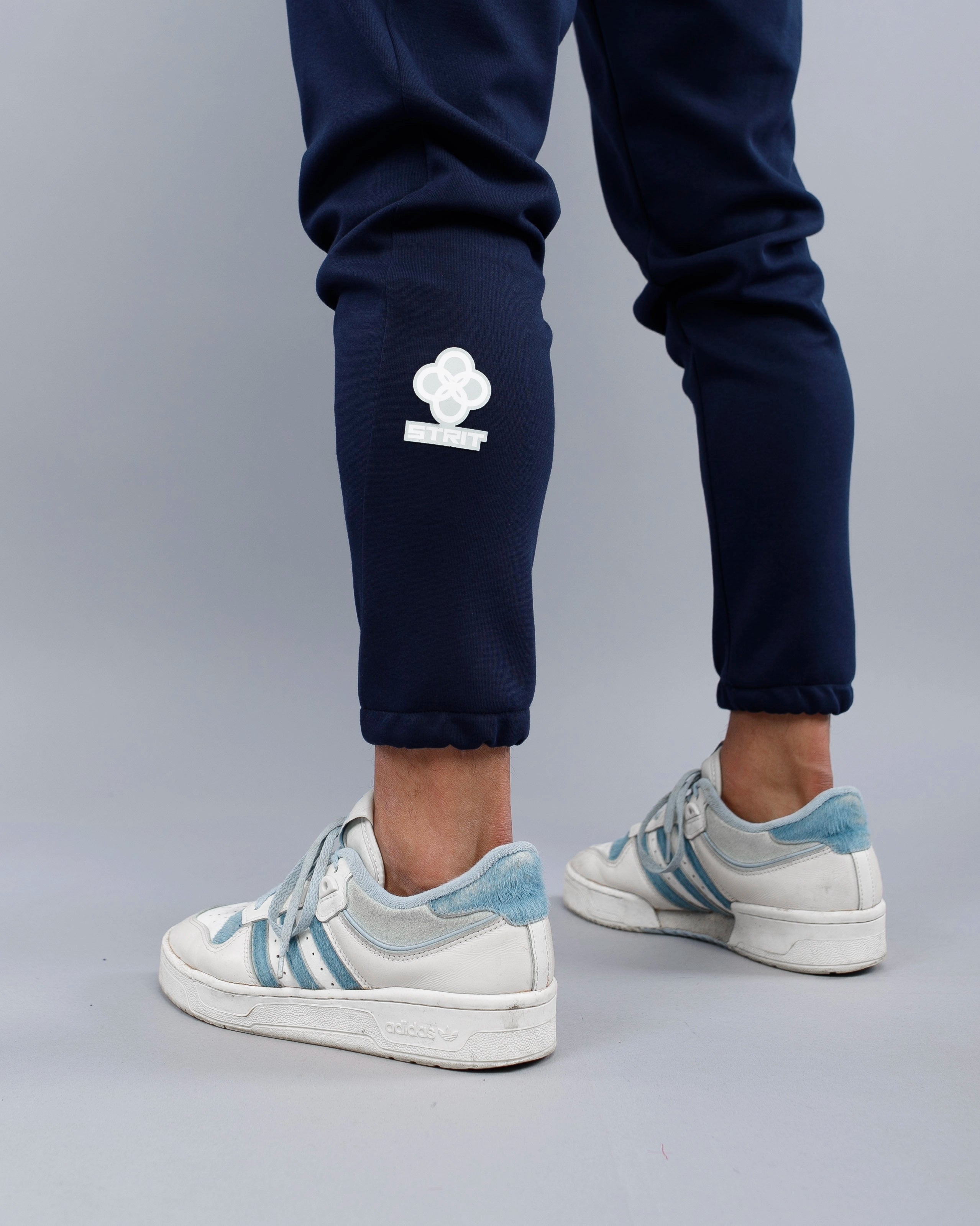 Navy blue jogger pants with a white logo worn by a person, paired with white Adidas sneakers on a gray background.