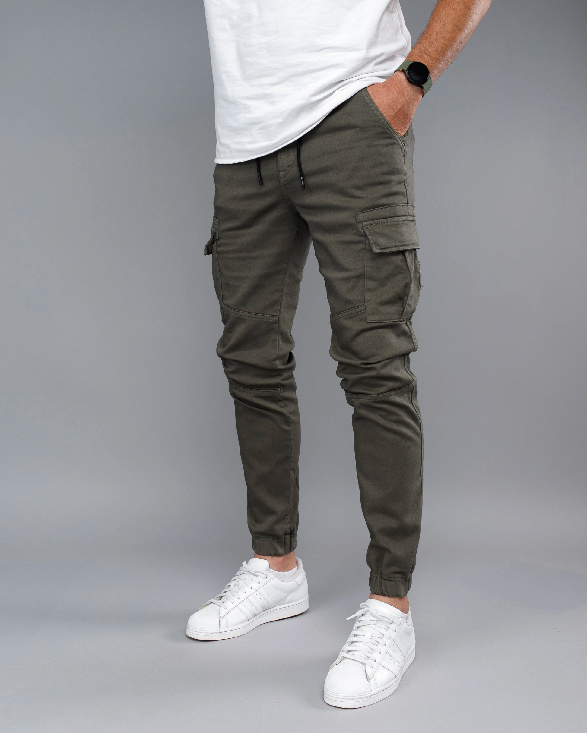 Person wearing olive green cargo jogger pants with a white t-shirt and white sneakers on a gray background