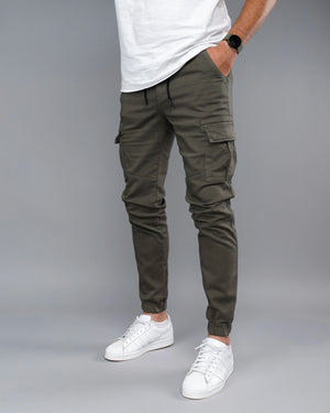 Person wearing olive green cargo jogger pants with a white t-shirt and white sneakers on a gray background
