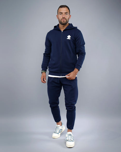 Man wearing a navy blue tracksuit with a logo on a gray background
