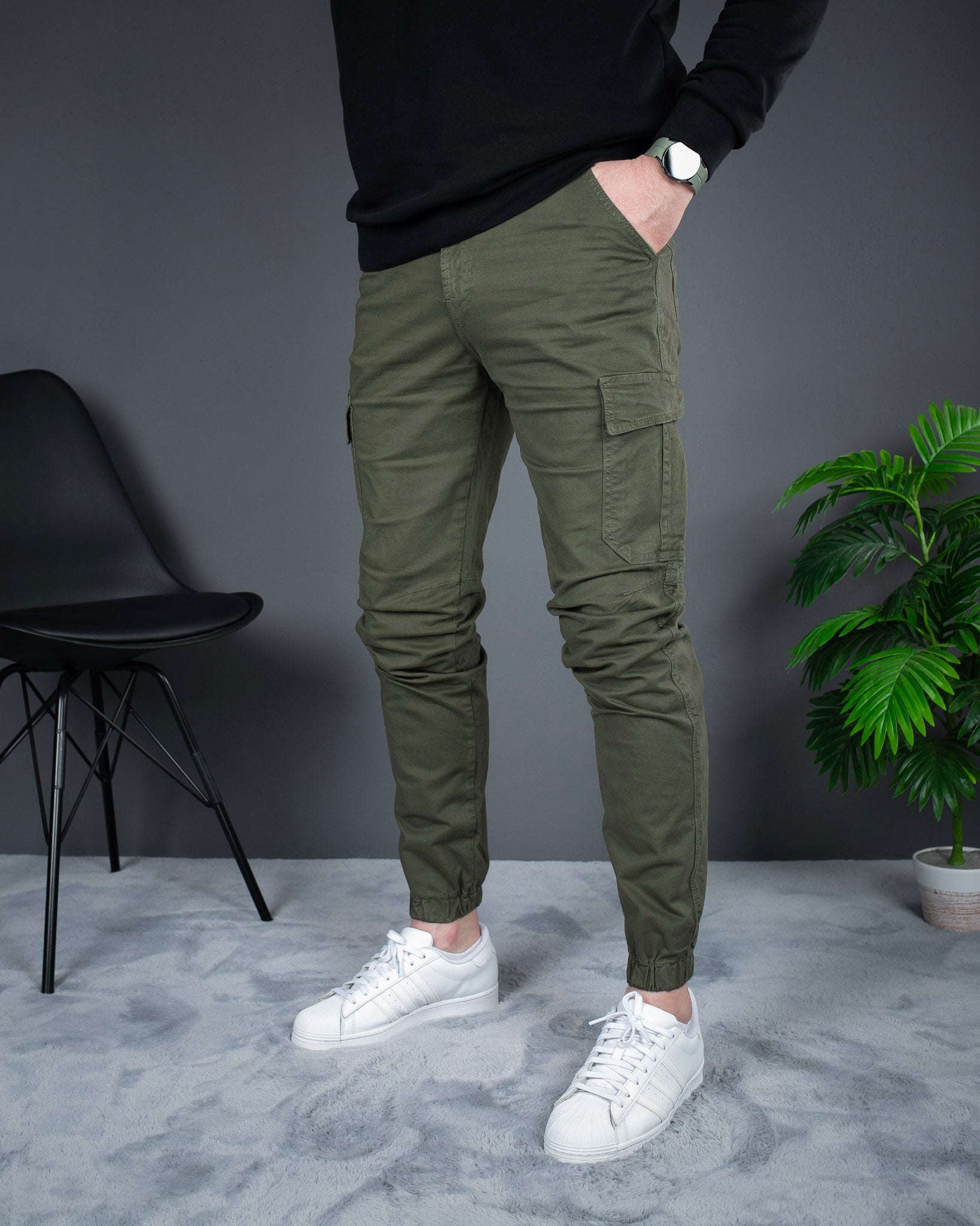 Person wearing olive green cargo pants and white sneakers in a room with a gray wall, black chair, and potted plant.